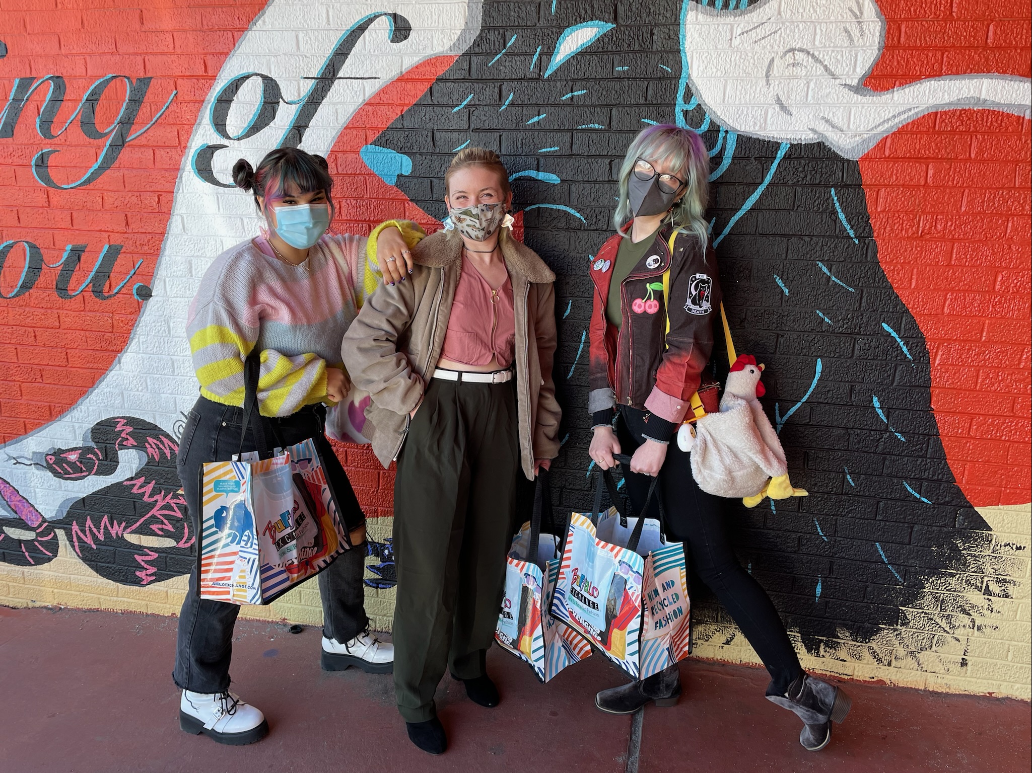 Three shoppers standing next to mural while holding Buffalo Exchange tote bags