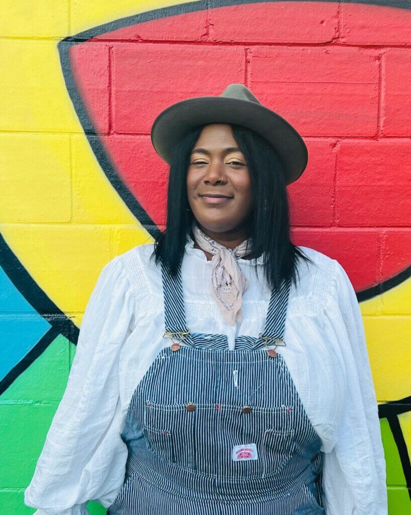 Person wearing a hat and dungarees stood in front of a brightly colored wall