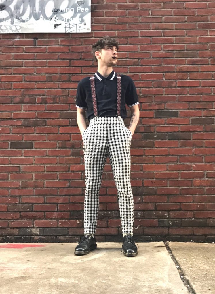 Man standing inside in front of shoe racks with foot up on bench wearing a yellow polo tee and green checkered socks