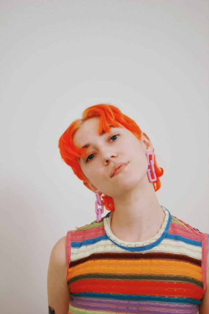 Person with orange hair, pink earrings, brightly colored striped top looking at camera