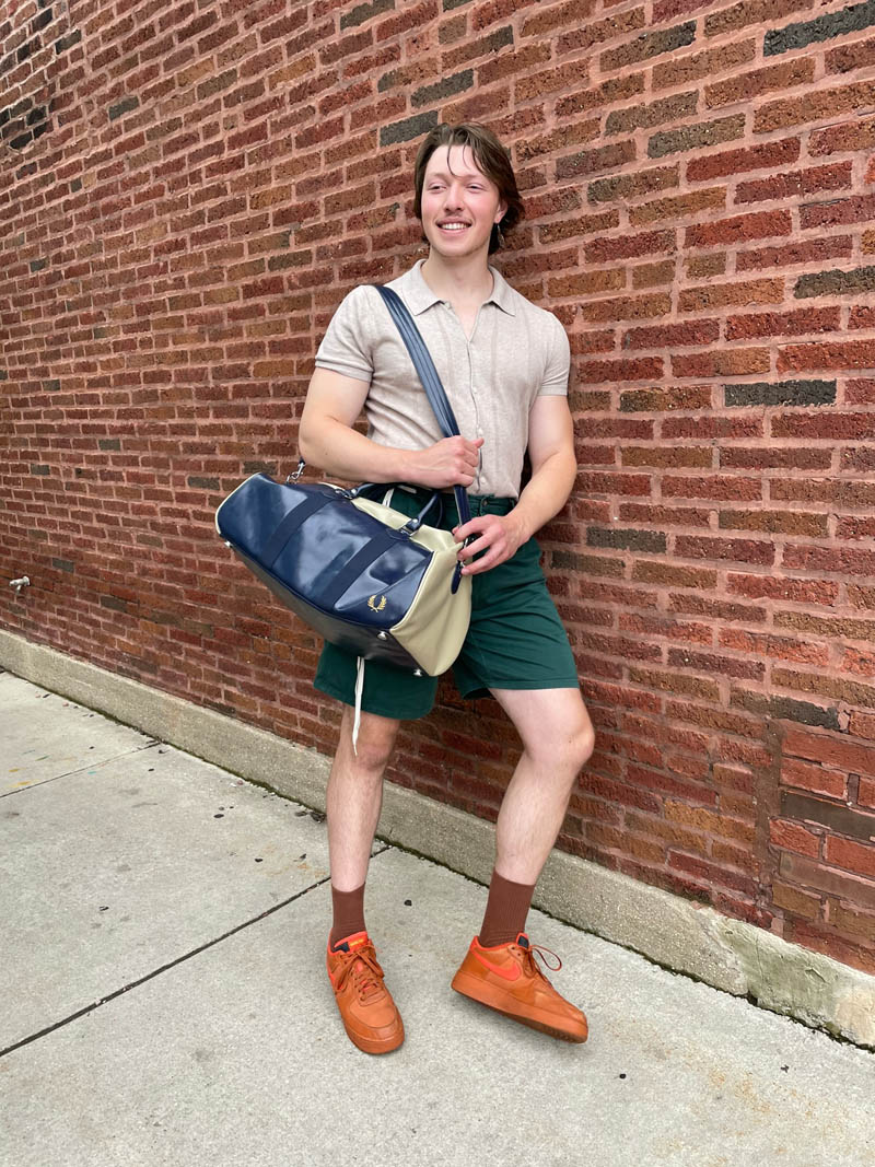 Person standing against brick wall wearing polo shirt, athletic shorts, Nike sneakers and holding duffel bag