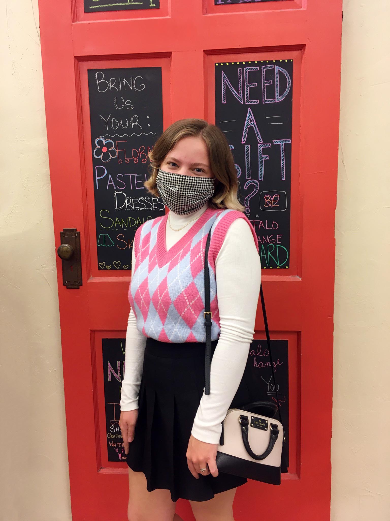 Person wearing face mask, argyle sweater vest styled with turtleneck underneath and pleated skirt with a Kate Spade crossbody bag on her shoulder