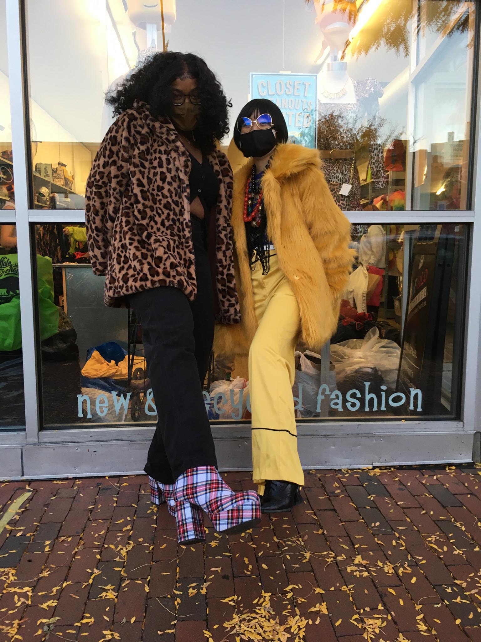 Two people posing in front of a store, the person on the right is wearing an animal print coat and the person on the right is wearing a yellow faux fur coat