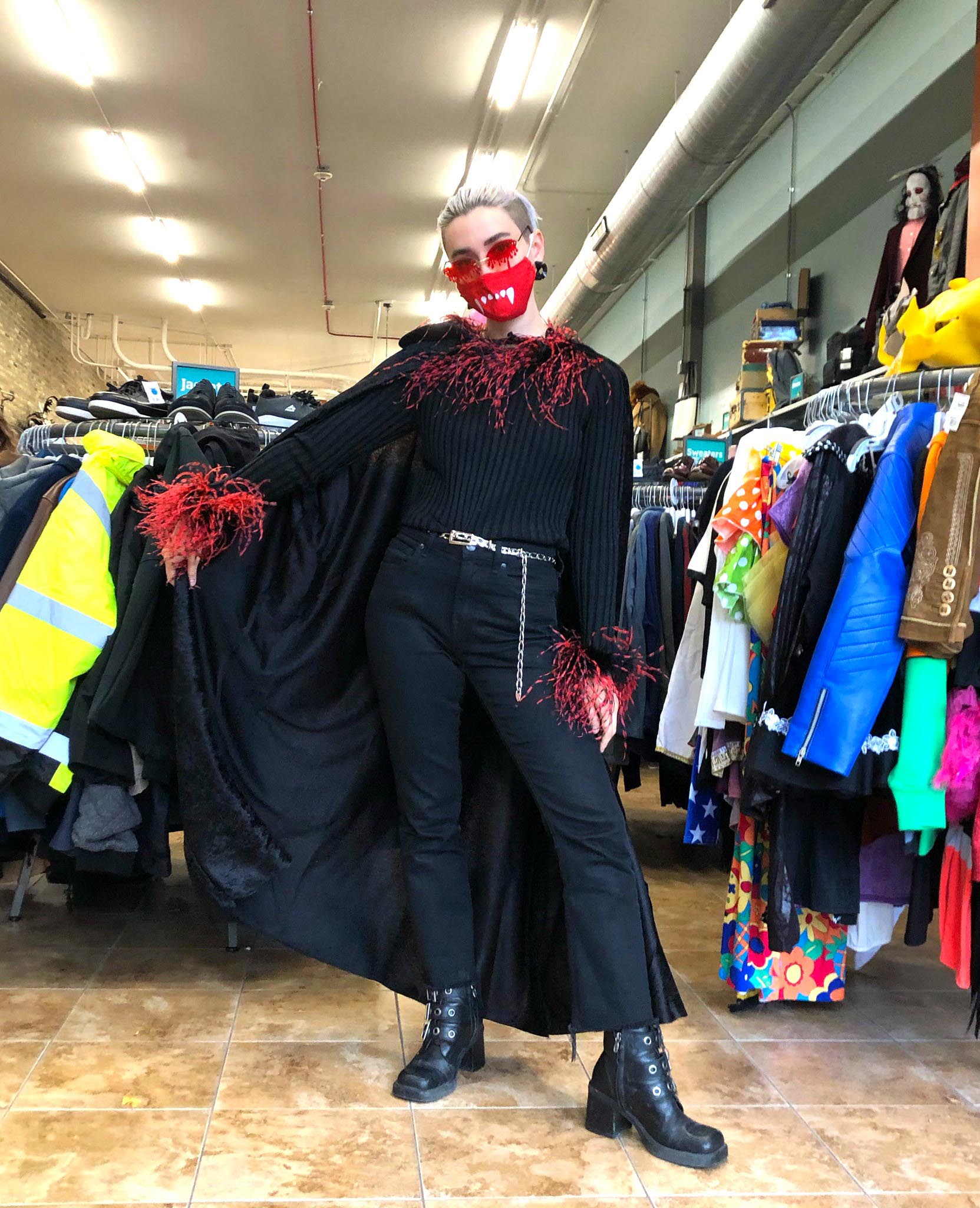 Person standing inside Buffalo Exchange store in front of clothing rounders wearing vampire costume with fang face mask, blood drip glasses and cape