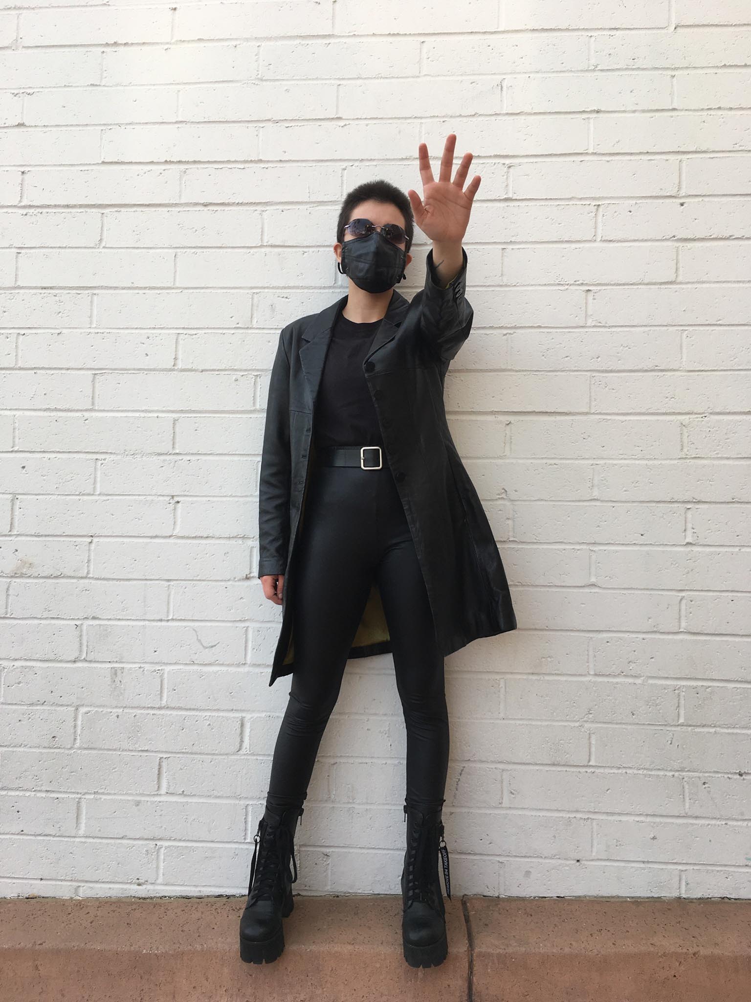 Person standing in front of brick wall in all black Matrix-inspired outfit with hand up in a stop motion