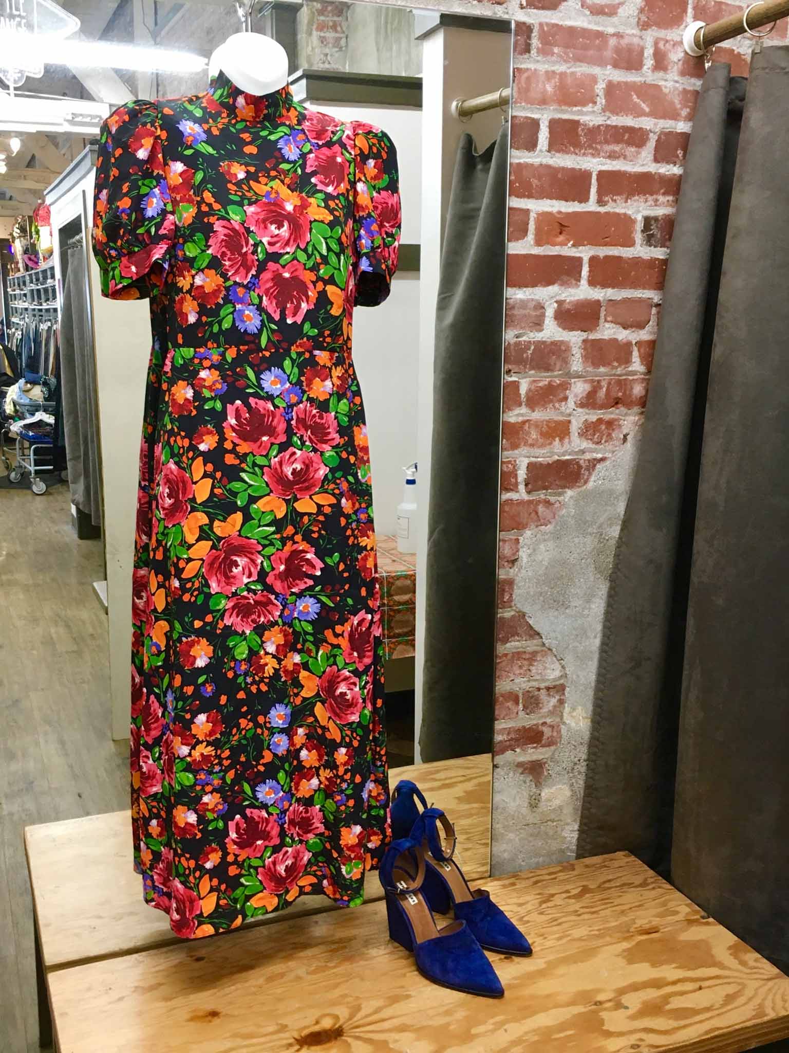 Floral midi dress and blue suede heels on mannequin hanging in front of wall-mounted mirror above wooden bench.