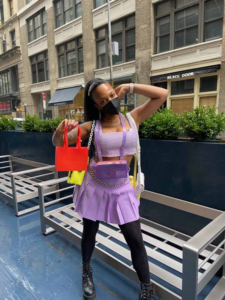 person poses in front of benches wearing lavender pleated skirt and matching top, holding 5 crossbody bags