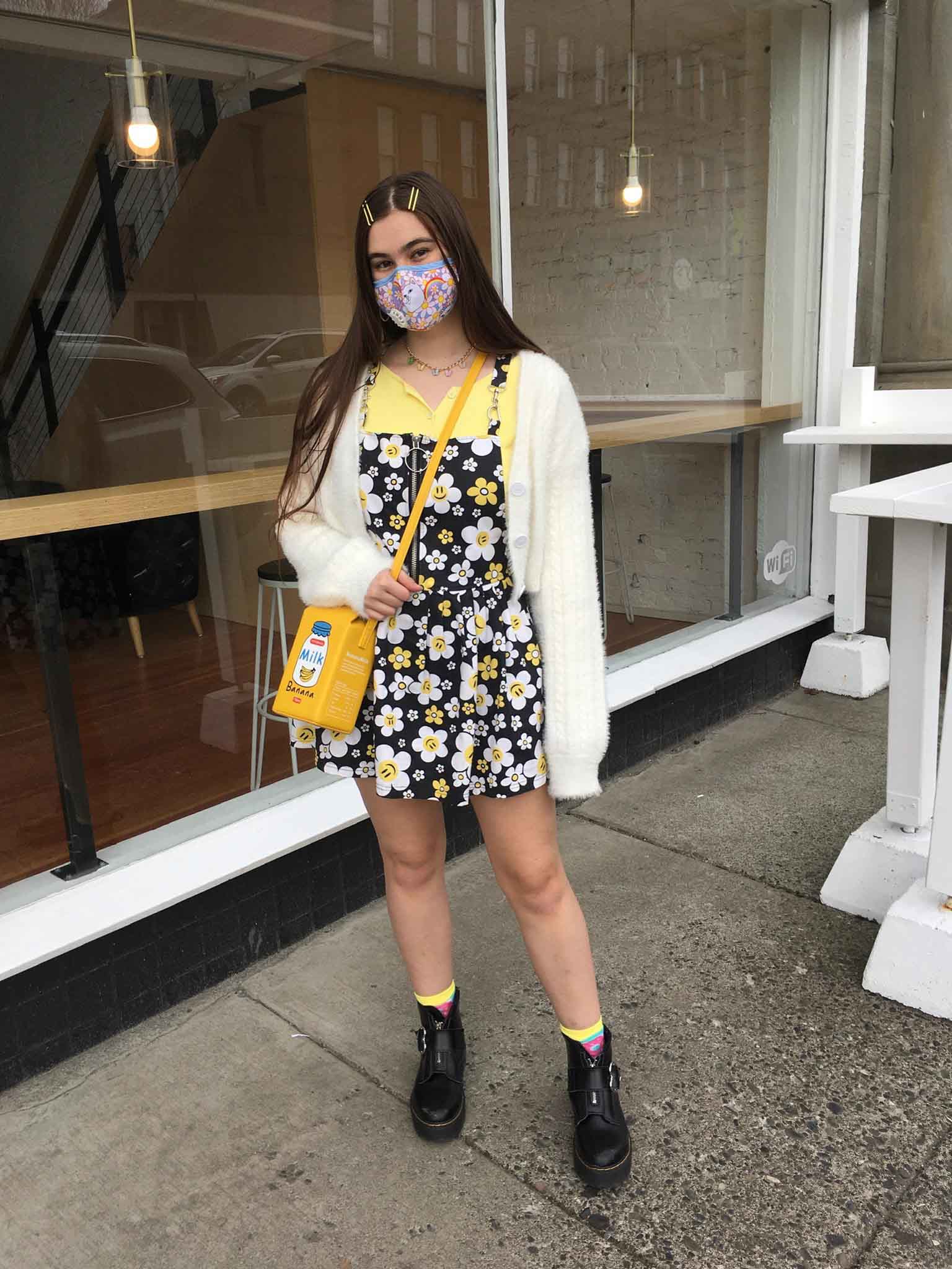 Person standing outside wearing large-print floral dress layered over yellow t-shirt, topped with cream cropped cardigan and black lace-up boots. Her novelty handbag is designed to look like milk carton.