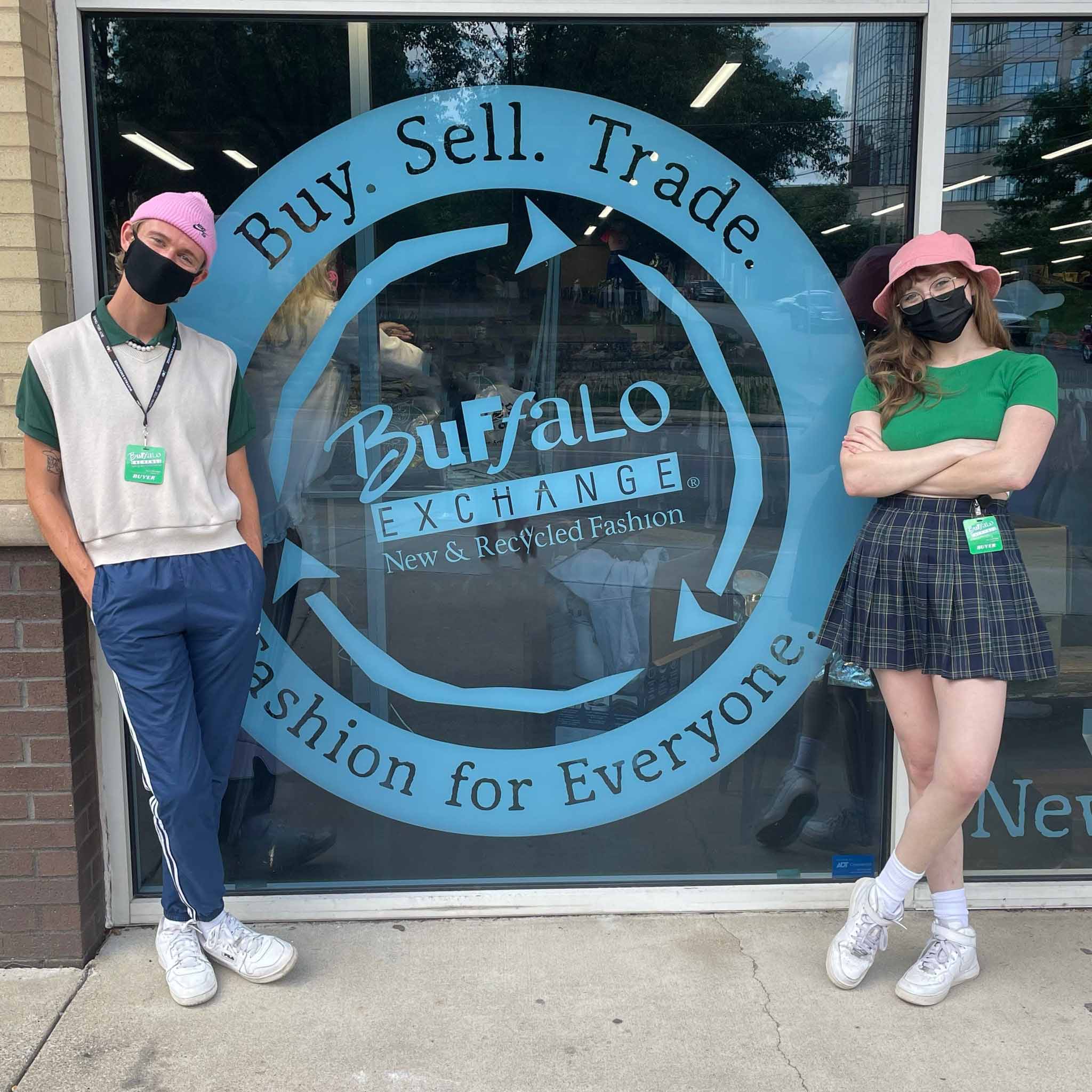 Two people stand in front of Buy.Sell.Trade sign at Buffalo Exchange Nashville. One person wears a sweater vest styled with t-shirt underneath, trackpants and beanie. Plaid pleated skirt styled with cropped green t-shirt and bucket hat, the other wearing pink bucket hat, cropped green tee shirt and plaid skirt