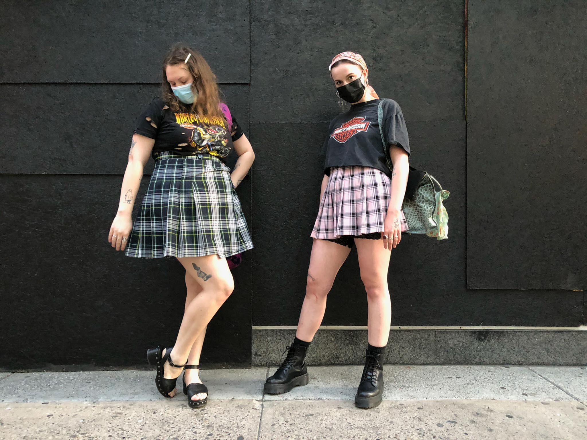 Two people stand on sidewalk in front of black wall wearing plaid skirts styled with vintage Harley Davidson t-shirts