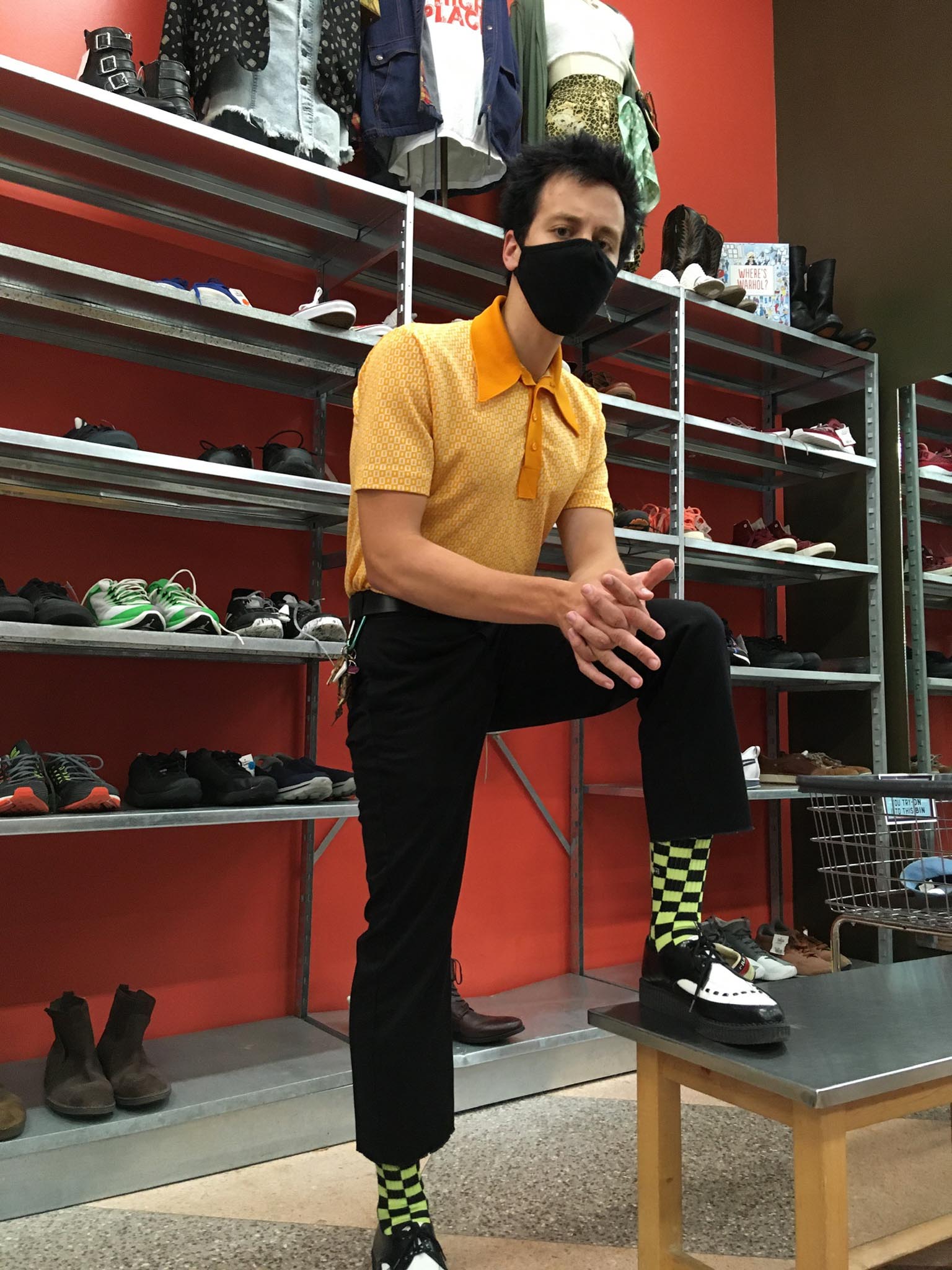Man standing inside in front of shoe racks with foot up on bench wearing a yellow polo tee and green checkered socks