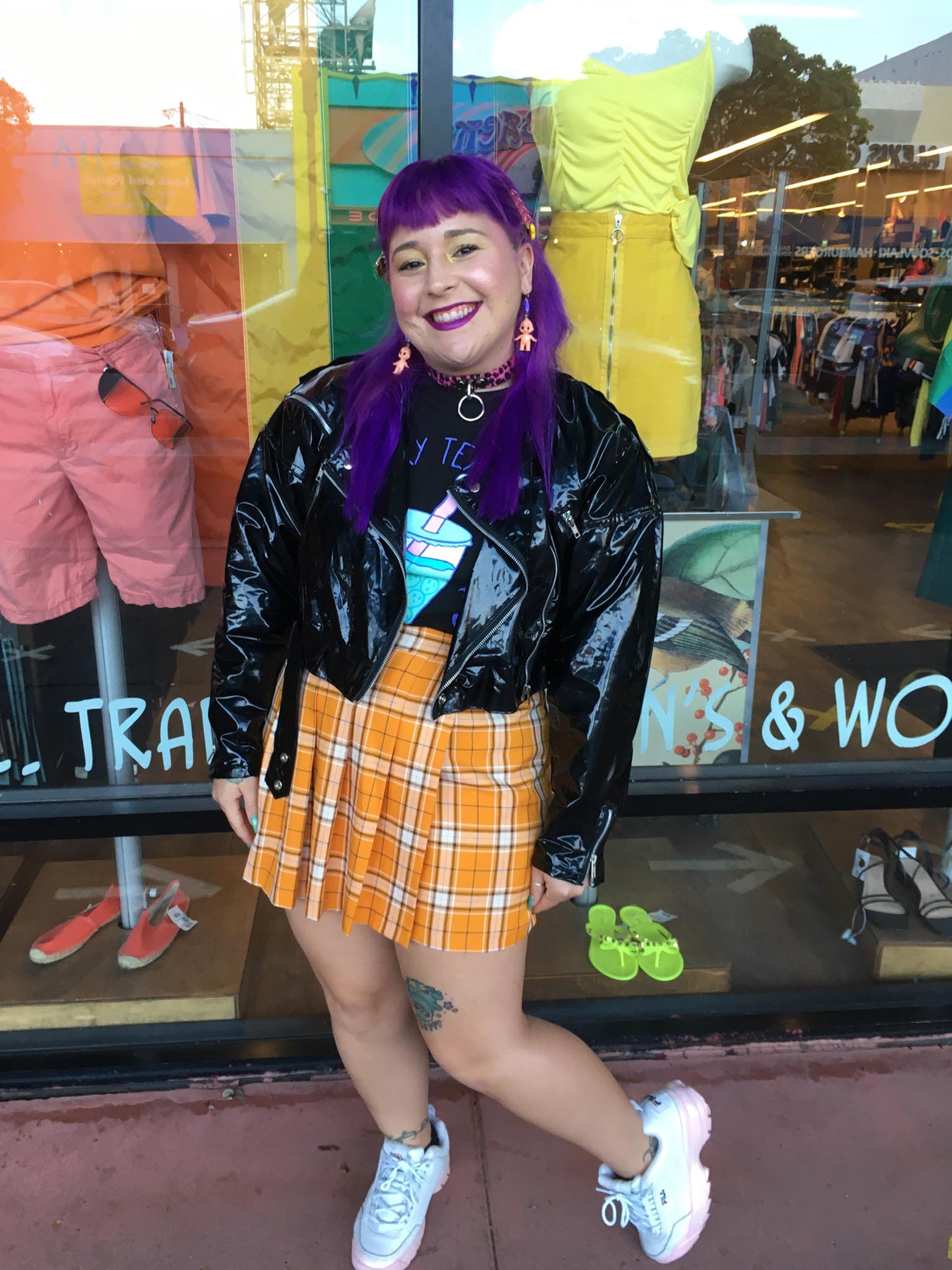 Smiling person with purple hair stands in front of shop window wearing an orange pleated skirt and black vinyl moto jacket