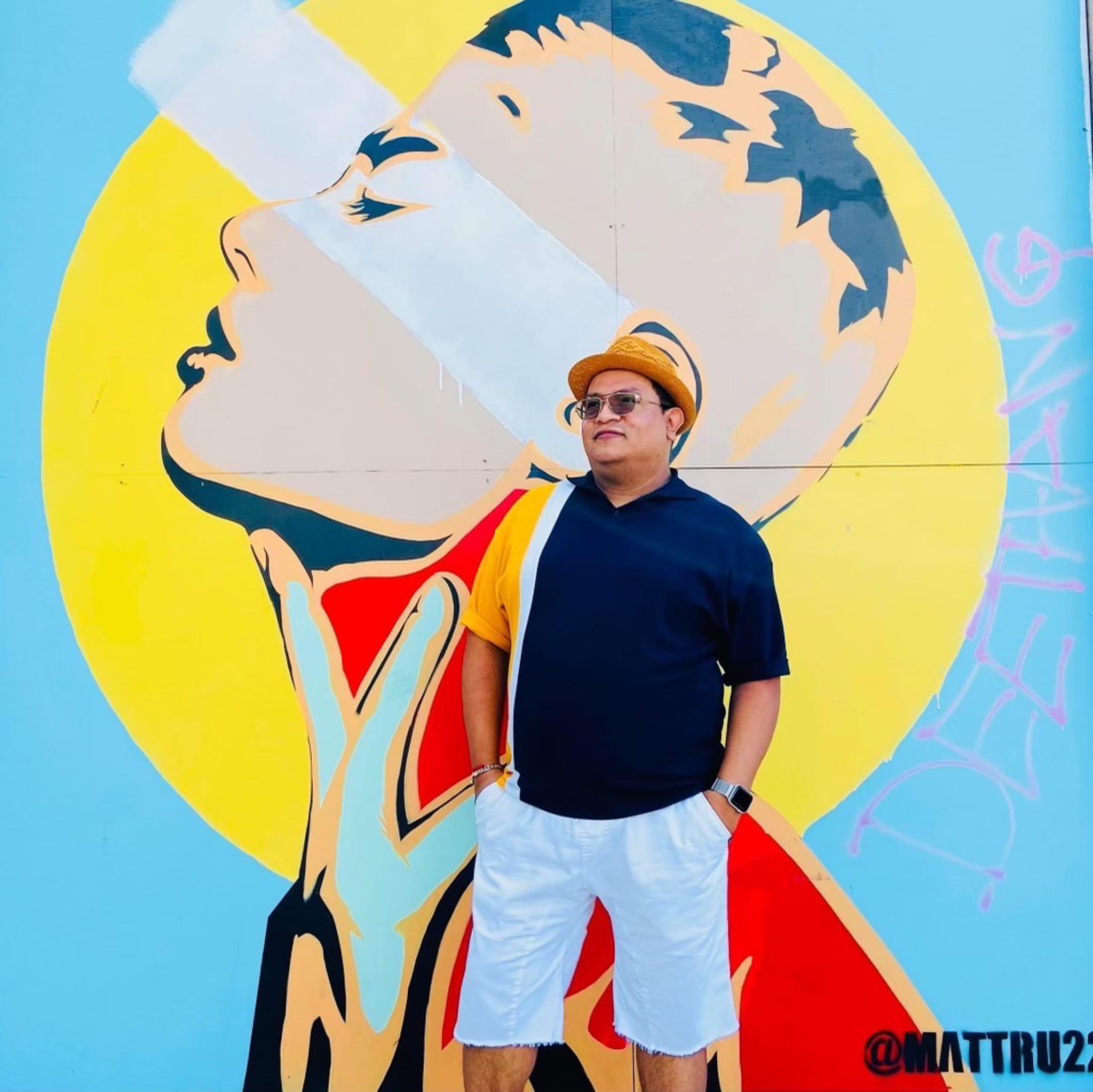 Felix posing in front of a colorful mural. He is wearing a blue, white and yellow polo t-shirt, white linen shorts and yellow fedora