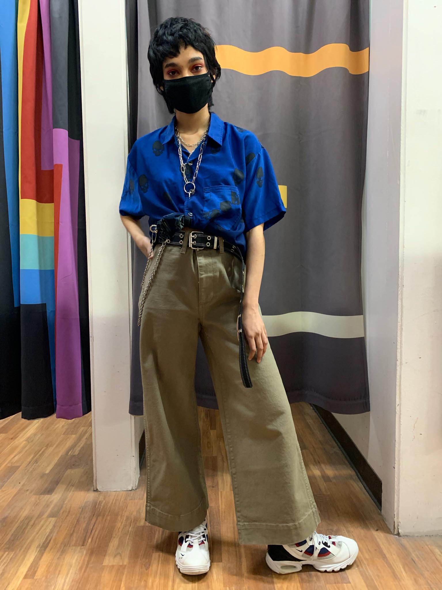 Person standing in front of Buffalo Exchange dressing room wearing royal blue short sleeved button down and wide leg khaki pants with metal jewelry and white Nike sneakers