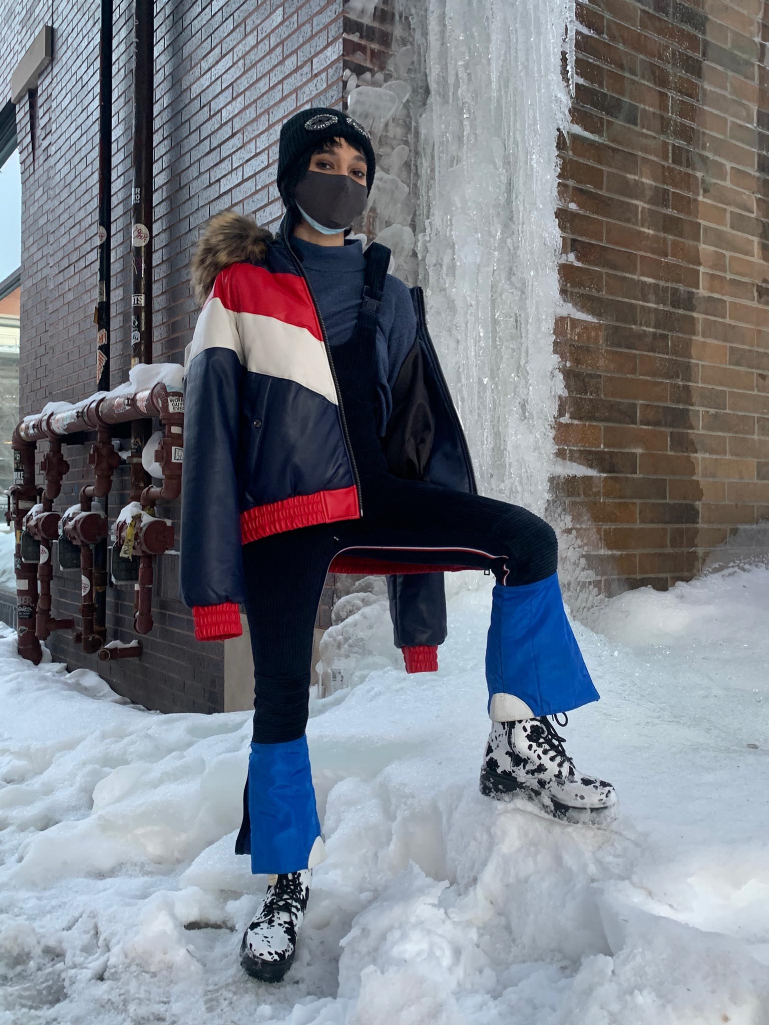A person stands on snow next to an iced-over building wearing vintage ski wear and cow print boots