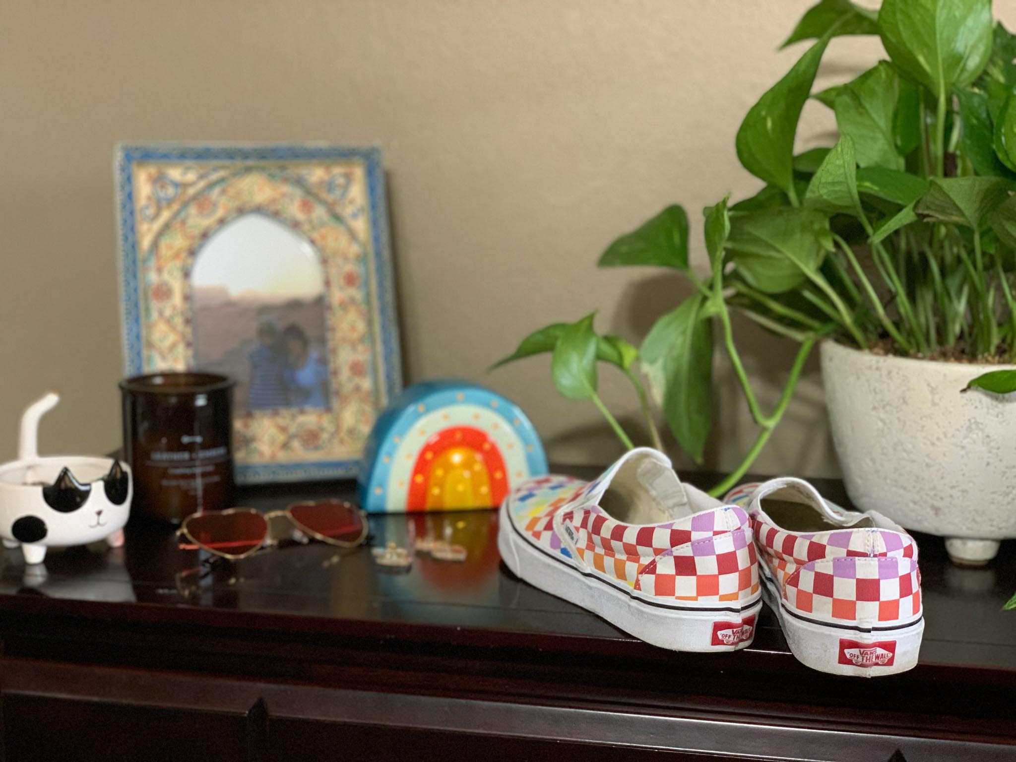 A pair of rainbow checker-print Vans on a wooden dresser, alongside a plant, sunglasses, picture frame, and candle