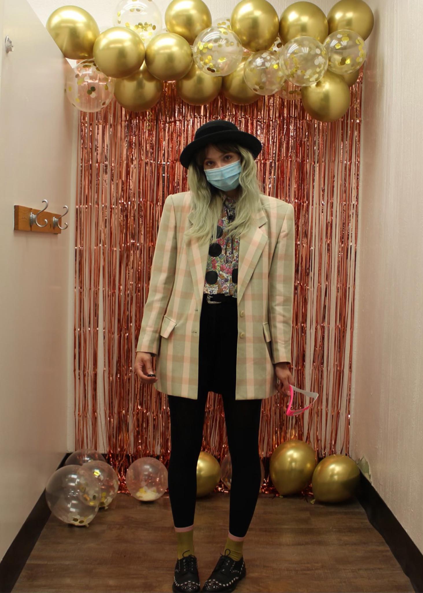 Woman standing in front of red tinsel and gold balloons wearing oversized plaid blazer, bowler hat and black skinny jeans
