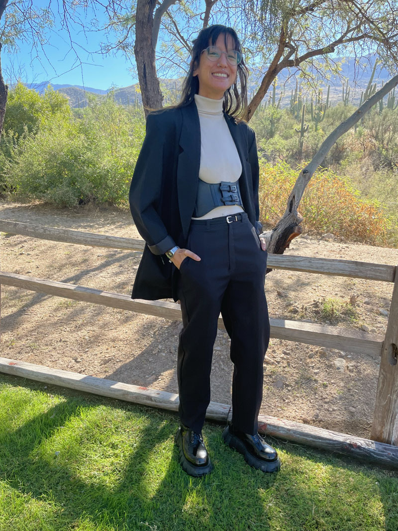 Photo of a smiling person dressed in semi-formal suiting while standing next to low wooden fence in field with desert background