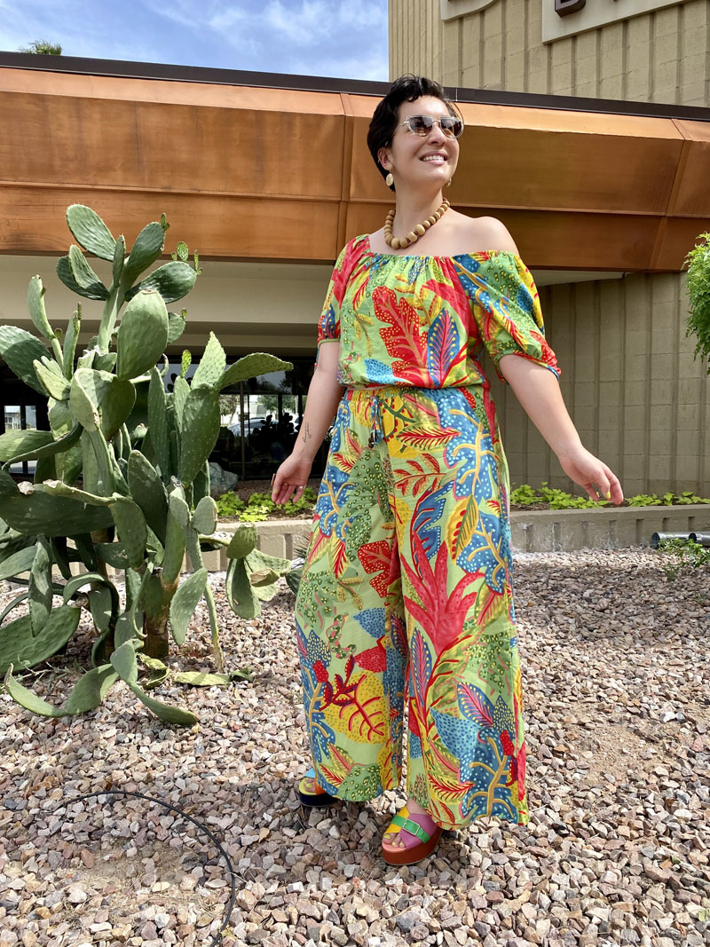 A person standing on a desert landscaped lawn in front of cactus wearing a brightly colored, tropical print jumpsuit