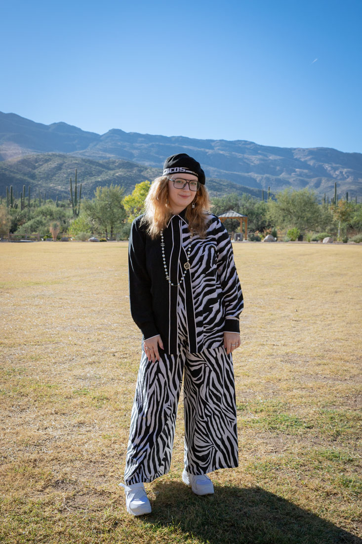 A person wearing a black beret, jacket and zebra-print jumpsuit while standing in an open field surrounded by mountains