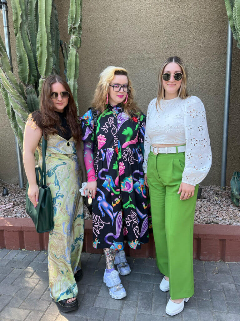 Three people posing together, the person on the left is wearing a green tones marbeling print dress and a green large purse, the person in the middle is wearing a bold print dress and lilac sneakers and the person on the right is wearing green trousers and a white eyelet blouse.