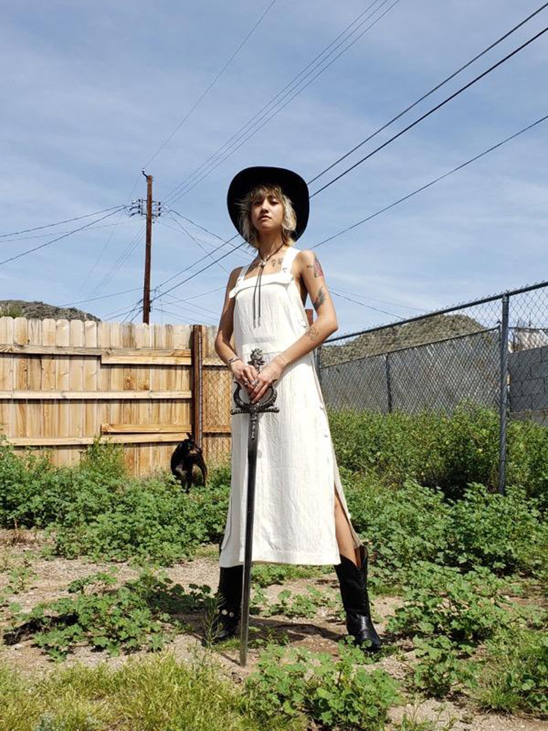 Person standing in fenced yard posed with a sword wearing white midi dress, black wide-brim hat and black boots
