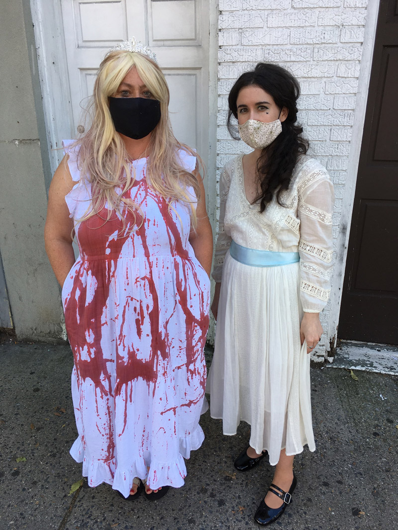 Two people in vintage gowns dressed like the characters Carrie and Baby Jane in long white vintage dresses. The person dressed as Carrie is wearing a tiara and their dress is splattered in fake blood.