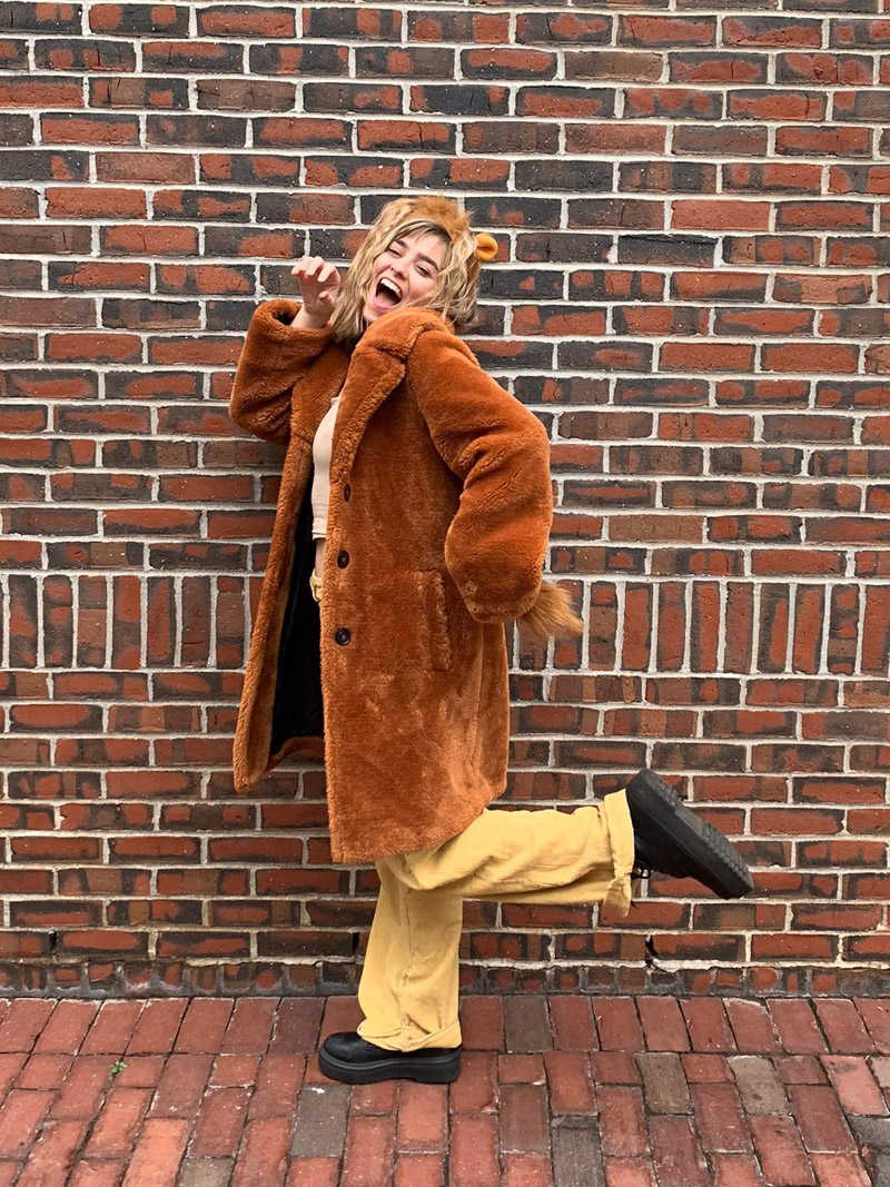 A person in a lion costume complete with lion ears, an oversized brown faux fur coat, yellow jeans, and black sneakers.