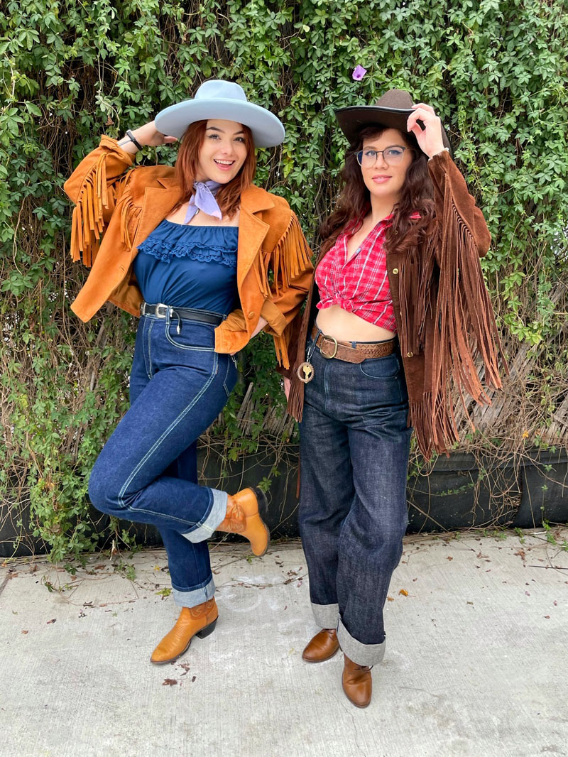 Two people dressed as cowboys in felt cowboy hats, fringe suede jackets, dark blue jeans with cuffed legs, and cowboy boots.