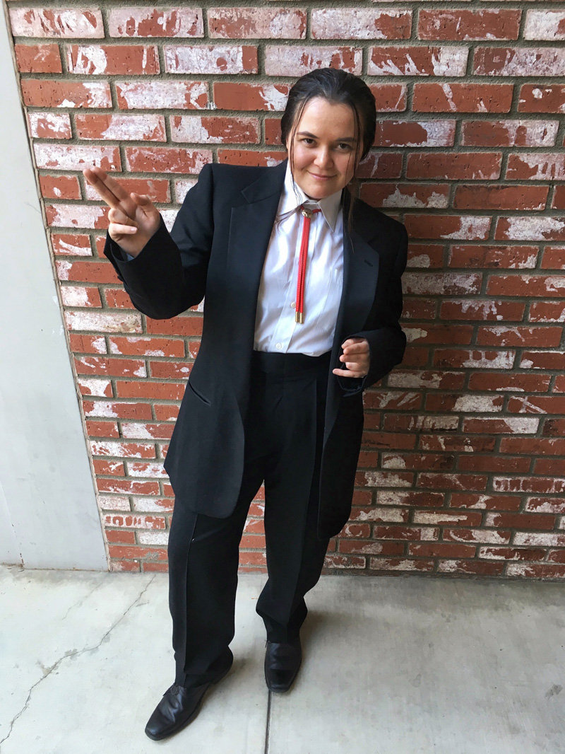 A person dressed as Vincent Vega from the movie Pulp Fiction, wearing a tuxedo, a bolo tie, and their hair pulled back in a ponytail.