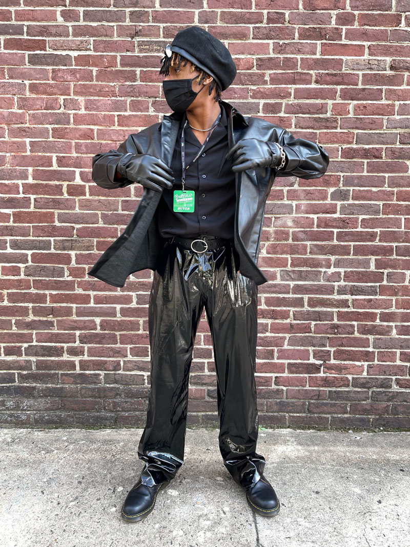 A person in a goth costume wearing head-to-toe black, including a newsboy cap, leather jacket, button down, baggy PVC pants, and Doc Martens boots.