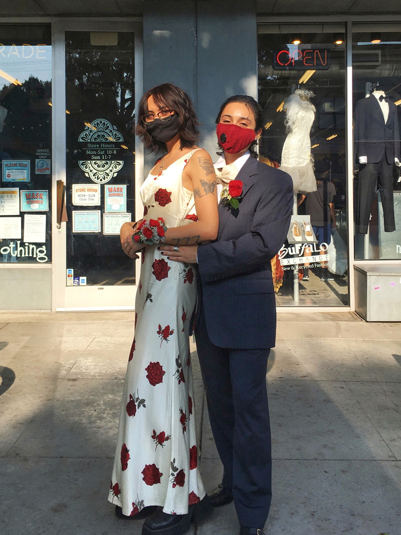 Two people dressed as a couple at prom, one person wearing a floral satin dress and corsage, the other in a gray tuxedo, an oversized white bowtie, and boutonniere.