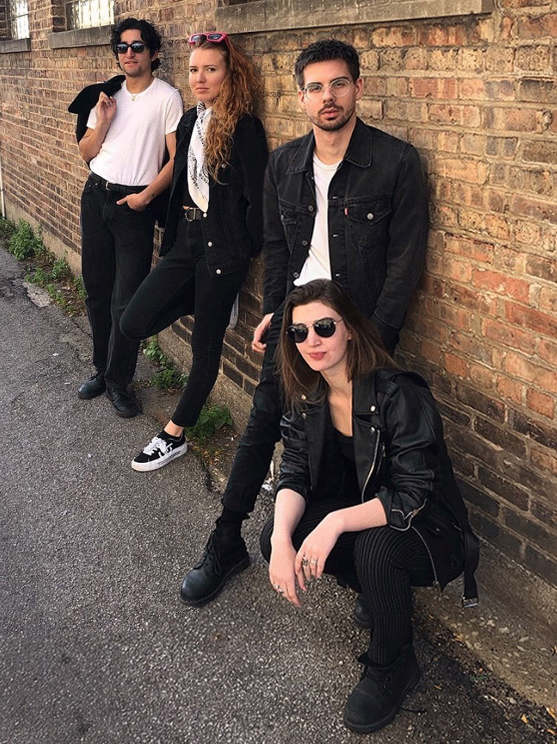A group of 4 people dressed as 50s Greasers in black and white outfits posing by an alley wall