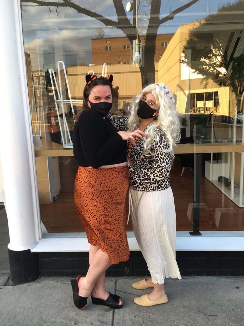 Two people dressed in simple wild cat costumes. One person is dressed as a tiger in a tiger ear headband, a black top, an orange and black spotted satin slip skirt. The other person is dressed as a leopard in a leopard print sweater, long cream-colored skirt, and nude shoes.