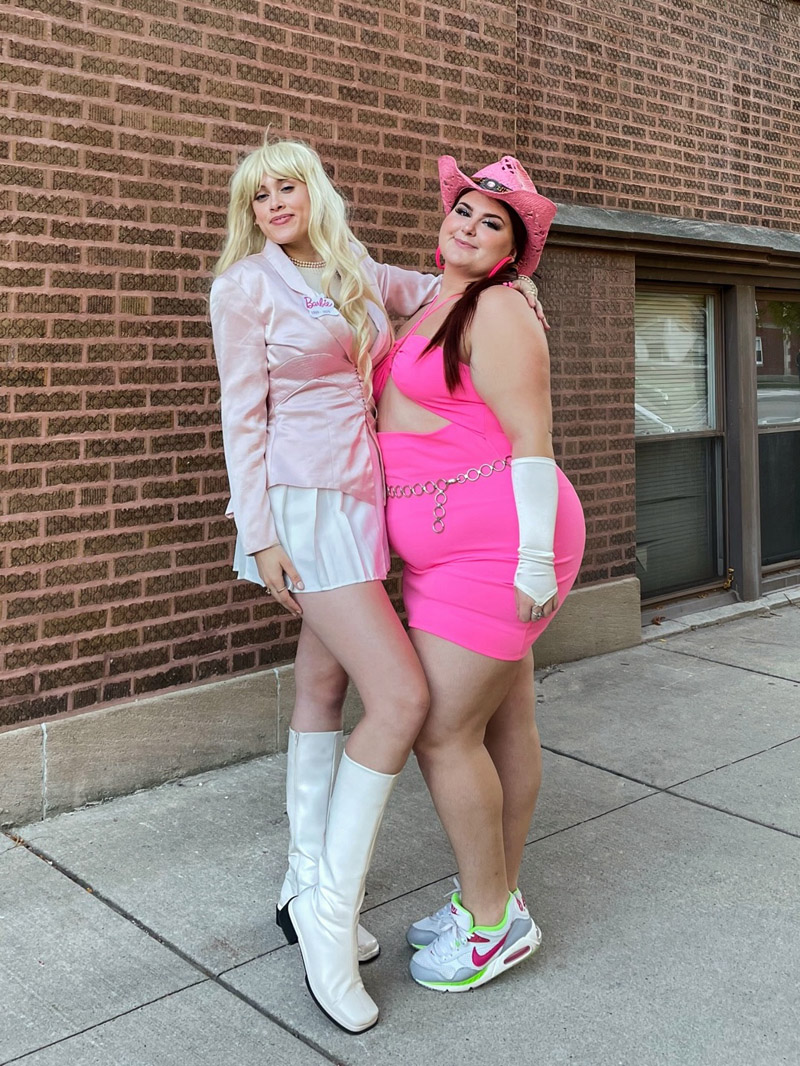 Two people in Barbie costumes, one person is wearing a light pink satin blazer, a white tennis skirt, and white knee-high boots, the other is wearing a pink cowboy hat, a hot pink mini dress, white arm sleeves, and chunky Nike sneakers.
