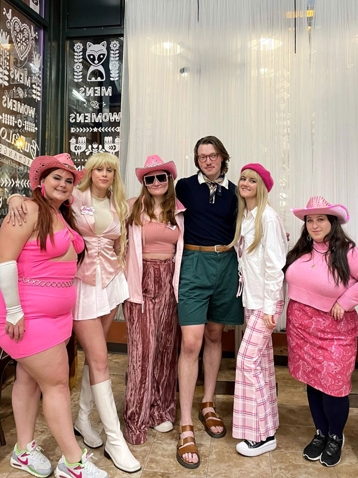 A group of people wearing different variations of Barbie costumes including cowgirl, preppy, and schoolgirl Barbie. A person dressed as Ken with a navy polo and green shorts stands in the middle.
