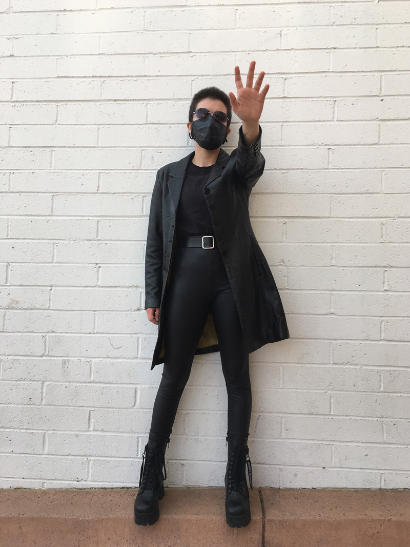 A person dressed up as Neo from the Matrix wearing sunglasses, a long black coat, a black top, black leggings, and black chunky platform boots.