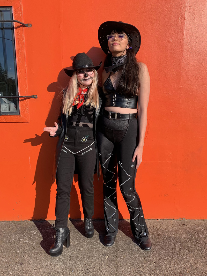 Two people wearing black cowboy hats, black tank tops, black jeans with western embellishments like embroidery or studs, and black boots.