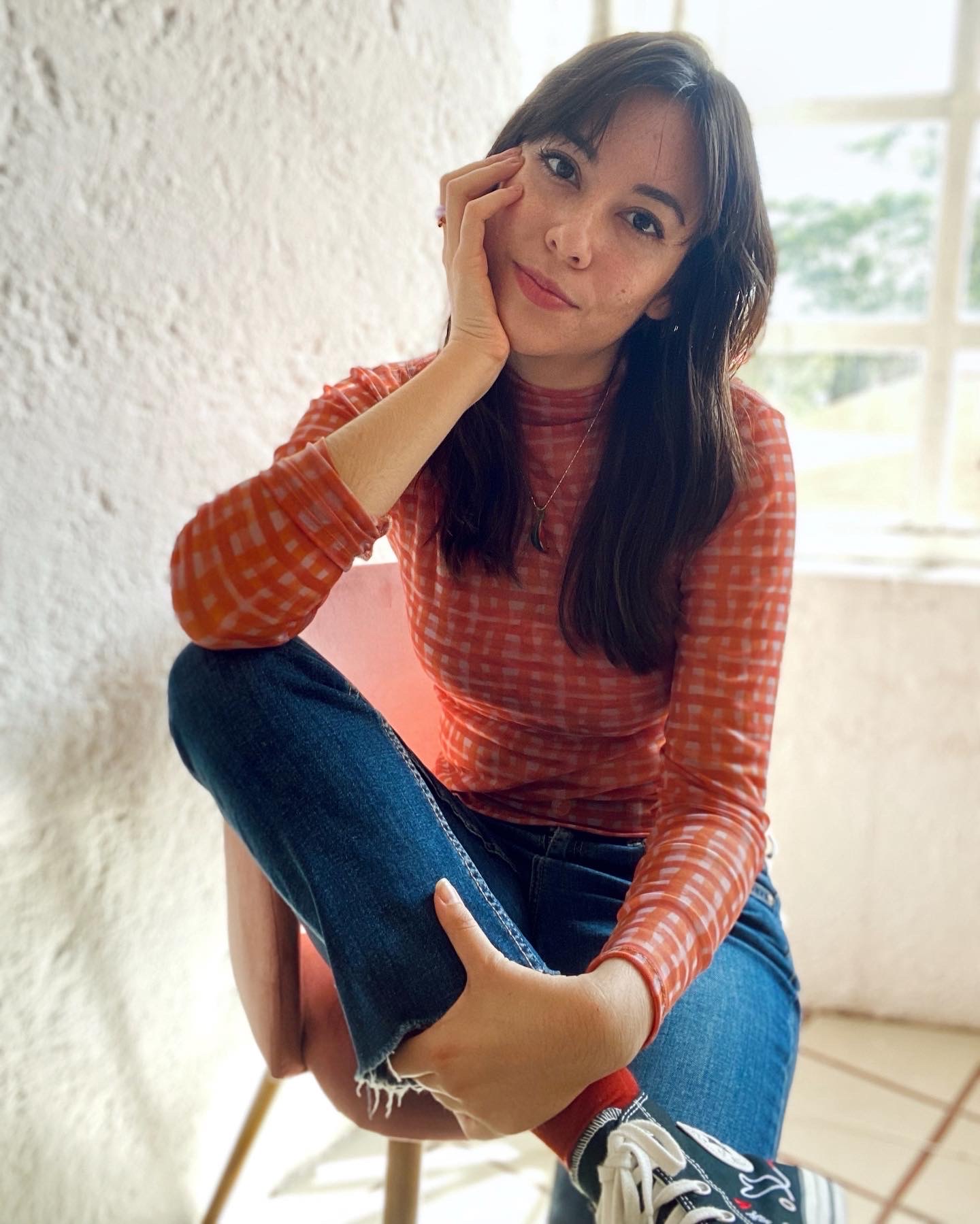 Girl posing sitting on a chair with a hand on her chin, she is wearinf blue jeans and a coral turtleneck t-shirt