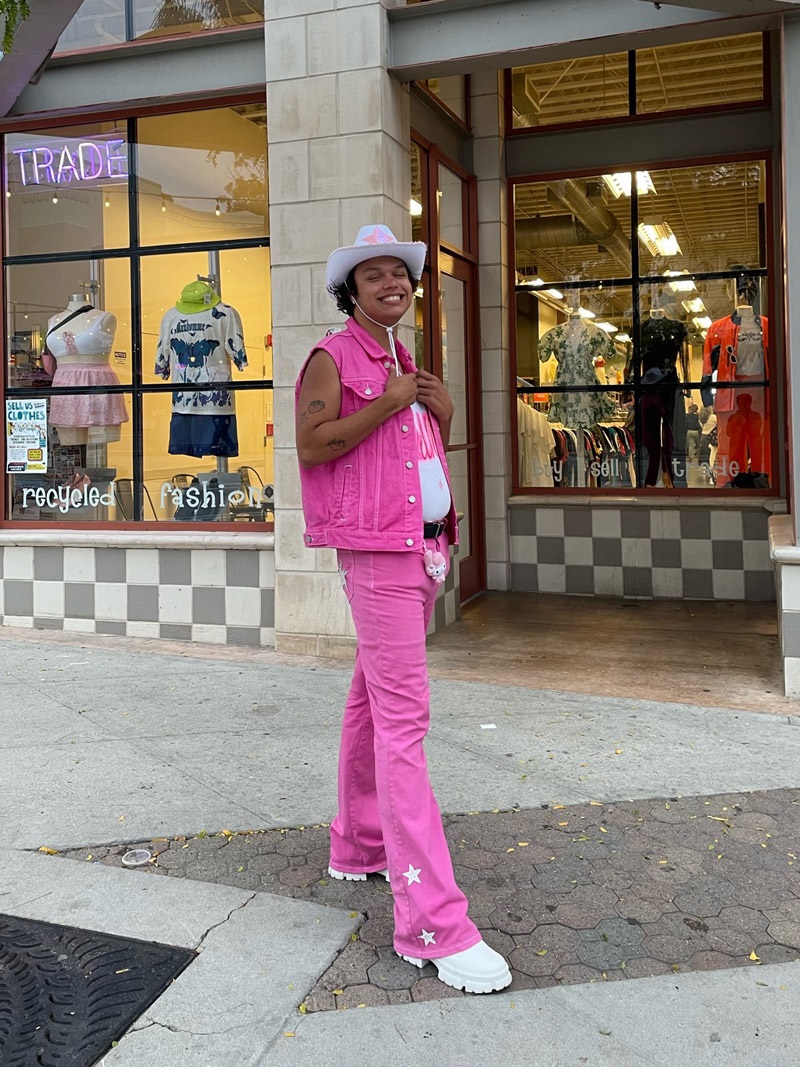 A person wearing a white cowboy hat, a pink denim vest, pink denim pants with white stars at the hem, and white lug sole boots.