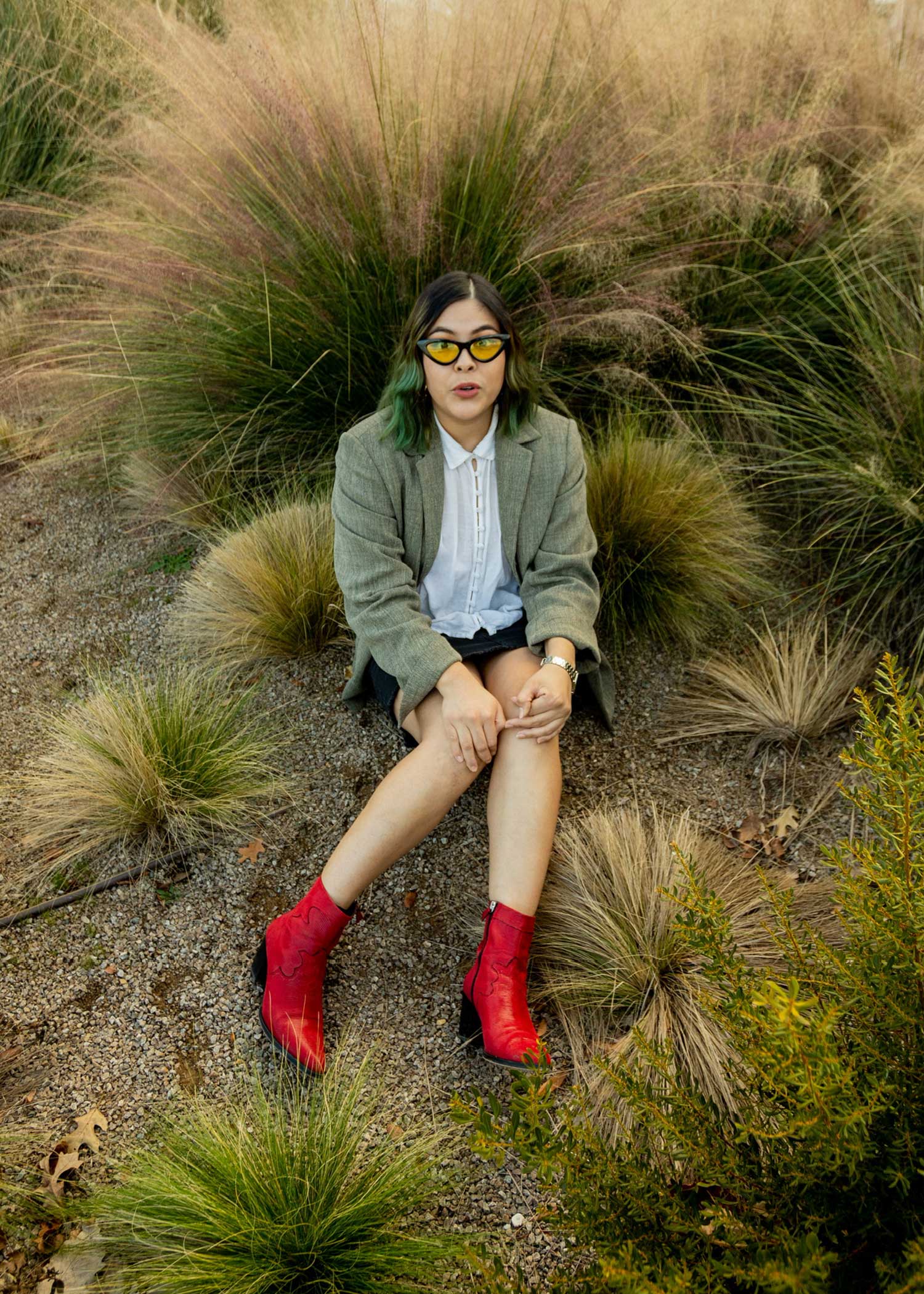 girl posing on the flow surrounded by desert plants, she is wearing bright red ankle boots, shorts, a white button up and a light green blazer