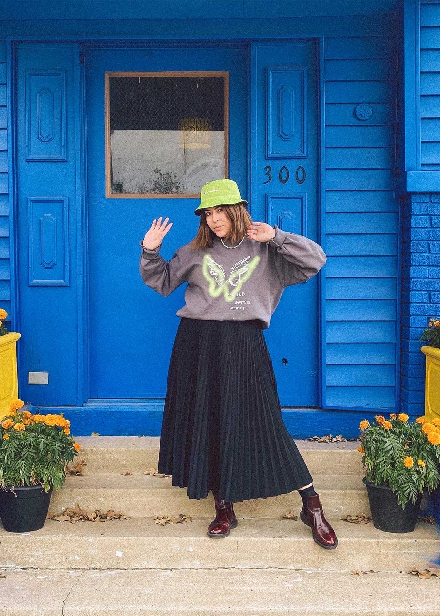 girl posing in front of a bright blue building wearing a maxi skirt, a sweatshirt and a bucket hat