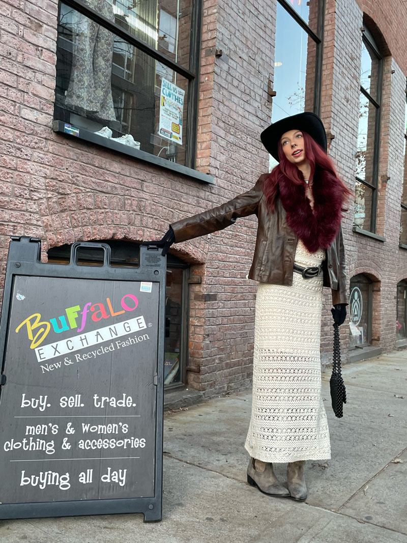 person wearing western hat, brown leather jacket and white crocheted dressstands on sidewalk holding onto Buffalo Exchange sandwich boar