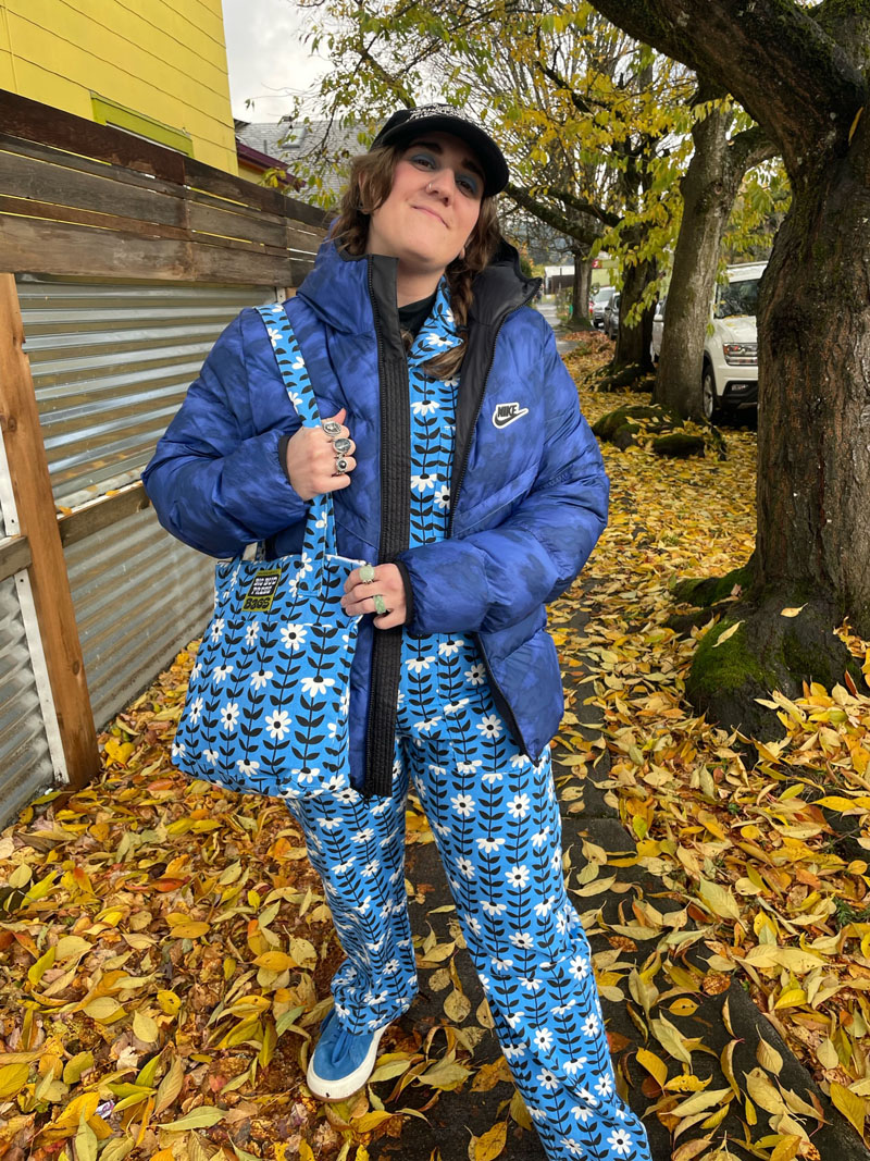 person standing on sidewalk covered in yellow leaves wearing floral jumpsuit with matching tote and blue puffer jacket 
