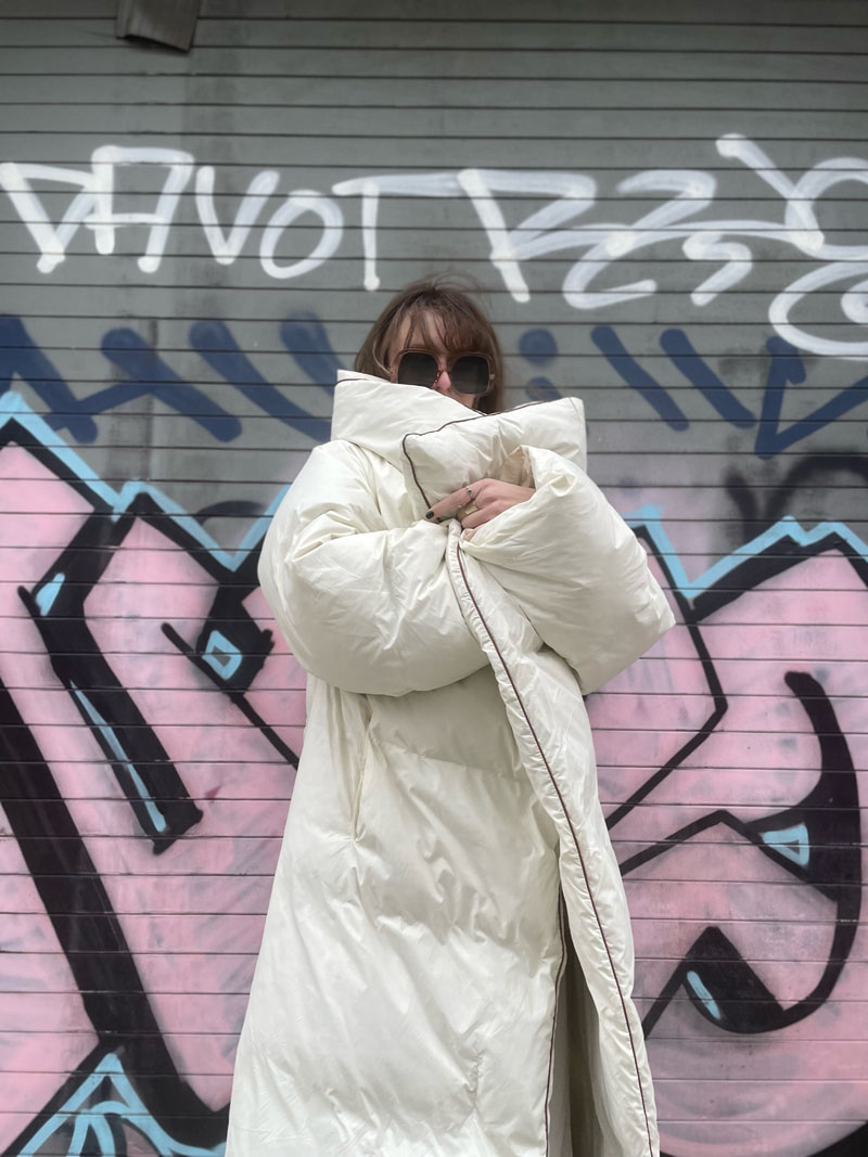 person standing in front of graffitied garage door wearing large white puffer jacket that partially conceals their face