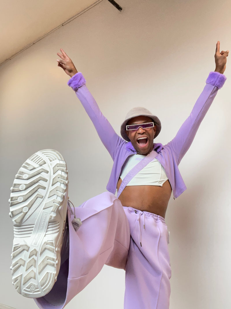 a person strikes a cheer pose while wearing lavender cropped sweater, pants, bucket hat and white tee shirt with sneakers
