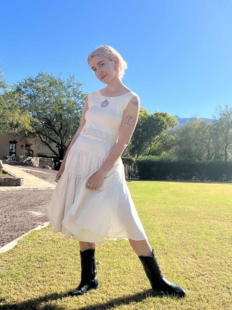 person wearing sleeveless white dress and black cowboy boots stands in a field with trees in the background