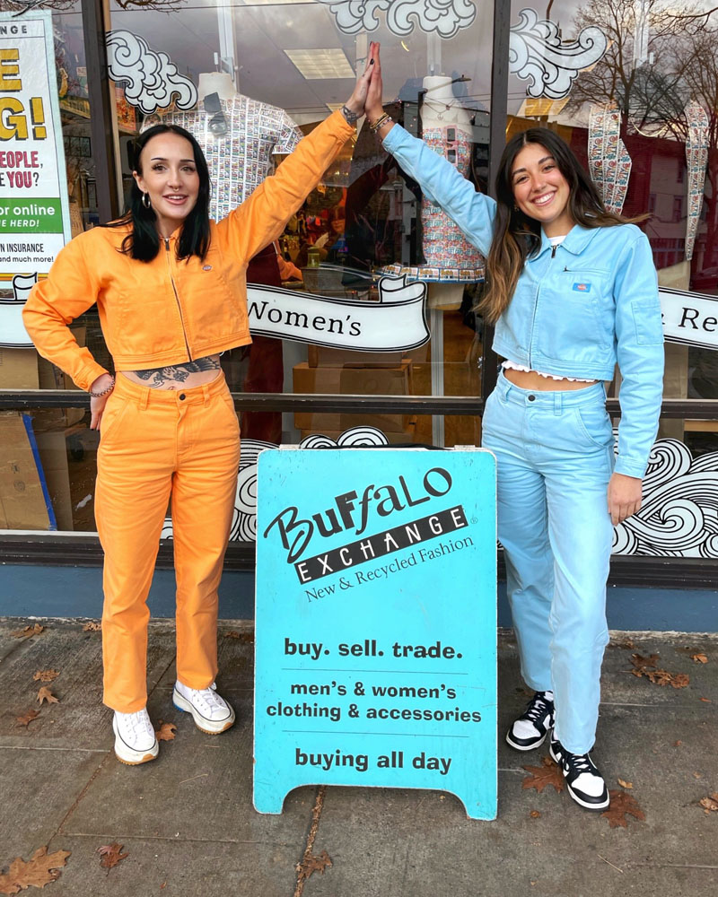 two people in coordinated Dickies two piece-sets high-five over teal Buffalo Exchange sandwich board between them 