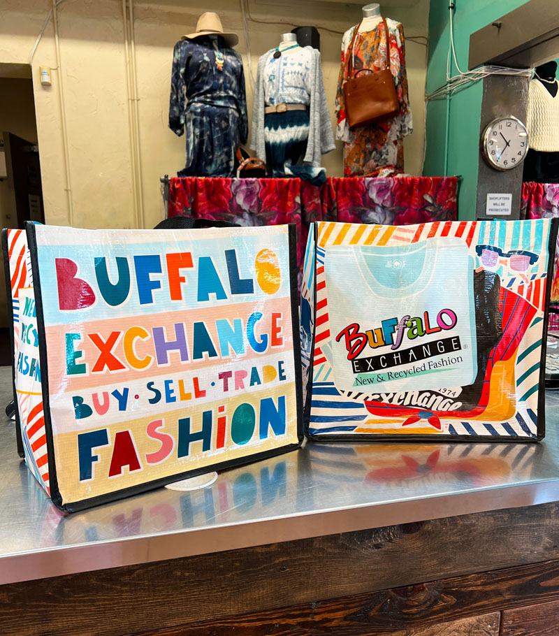 two colorful Buffalo Exchange tote bags sit on a metallic counter, with mannequins dressed in western outfits in the background