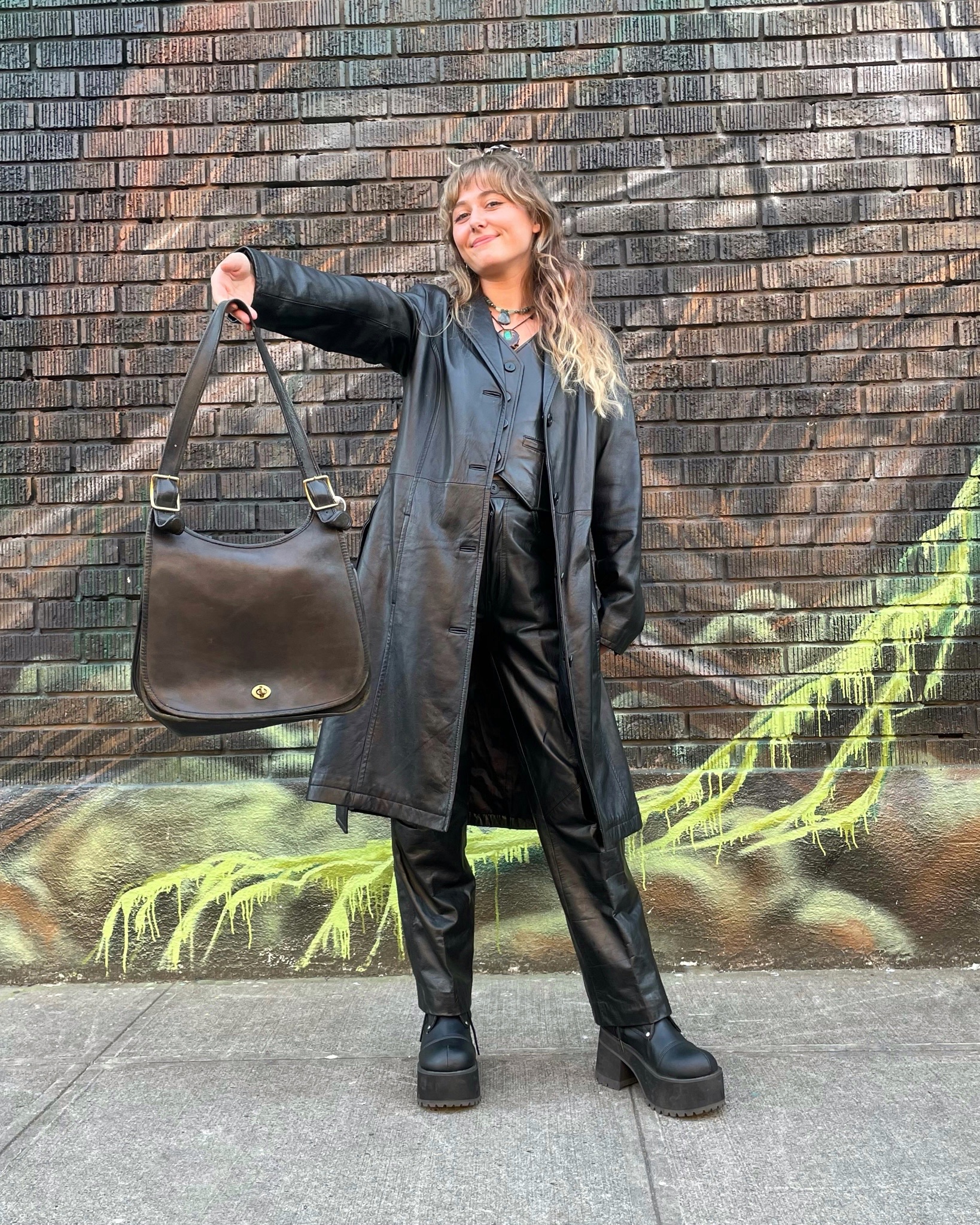 Woman posing in front of a brick wall wearing all black leather holding a black vintage Coach bag